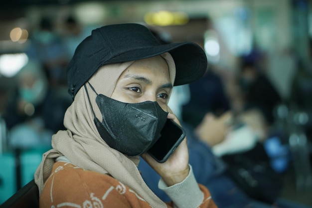 Sorridente giovane donna asiatica in hijab che indossa maschera e cappello seduto in sala d'attesa