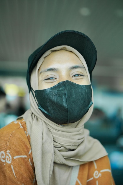 Sorridente giovane donna asiatica in hijab che indossa maschera e cappello seduto in sala d'attesa