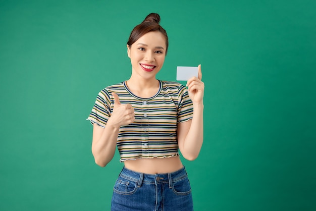 Sorridente giovane donna asiatica in piedi oltre il muro verde e tenendo la carta di debito nelle mani.