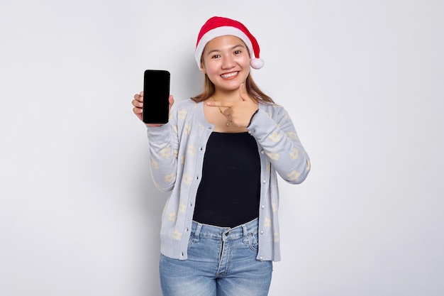 Sorridente giovane donna asiatica in un cappello di Natale che mostra il telefono cellulare con schermo vuoto che raccomanda l'app mobile isolata su sfondo bianco