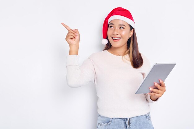 Sorridente giovane donna asiatica in un cappello di Natale con in mano una tavoletta digitale e puntando il dito da parte isolato su sfondo bianco Il popolo indonesiano celebra il concetto di Natale