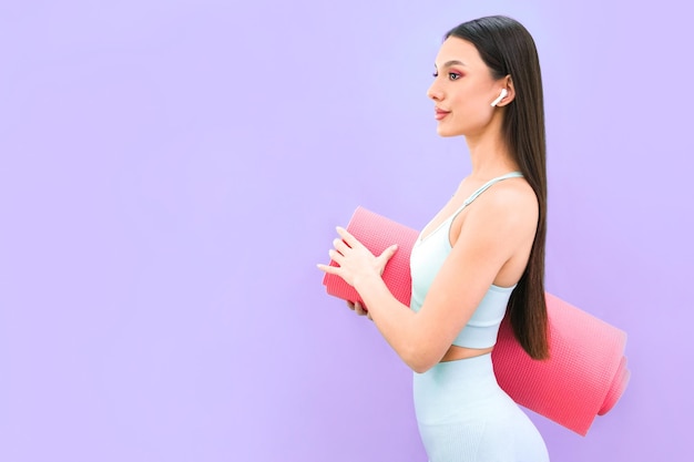 Sorridente giovane donna atletica in abbigliamento sportivo in posa contro un muro di studio viola Il concetto di allenamento per la motivazione sportiva Copia spazio Tenere un tappetino da yoga