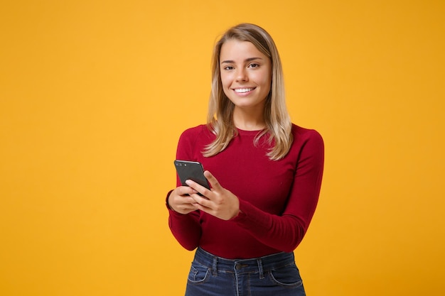 Sorridente giovane donna bionda ragazza in abbigliamento casual in posa isolato sul ritratto in studio di sfondo parete giallo arancio. Concetto di stile di vita delle persone. Simula lo spazio della copia. Utilizzando il telefono cellulare digitando un messaggio sms.