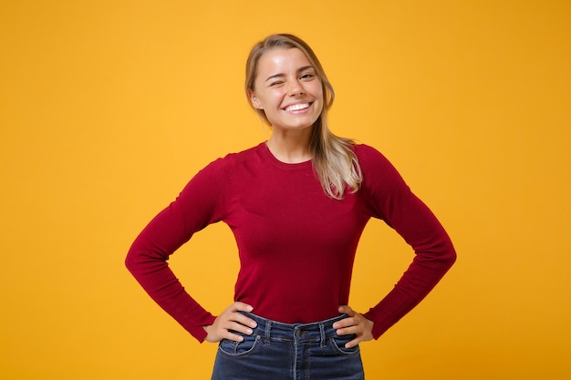 Sorridente giovane donna bionda ragazza in abiti casual in posa isolato su sfondo giallo arancione ritratto in studio. Concetto di stile di vita delle persone. Simula lo spazio della copia. Stare con le braccia sui fianchi in vita, lampeggiante.