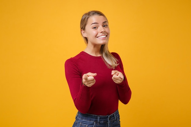 Sorridente giovane donna bionda ragazza in abiti casual in posa isolato su sfondo giallo arancione ritratto in studio. Concetto di stile di vita di emozioni della gente. Simula lo spazio della copia. Indice puntato sulla fotocamera.