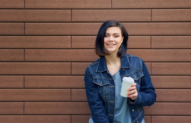 Sorridente giovane donna bruna con tunnel nelle orecchie in una giacca di jeans in piedi davanti al muro di mattoni godendo nel suo caffè caldo.
