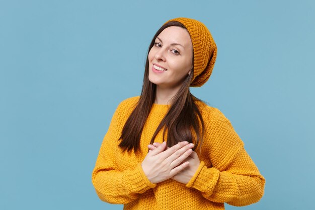 Sorridente giovane donna bruna in maglione giallo cappello in posa isolato su sfondo blu parete ritratto in studio. Concetto di stile di vita delle emozioni delle persone. Mock up spazio di copia. Tenendosi per mano piegata sul petto, cuore.