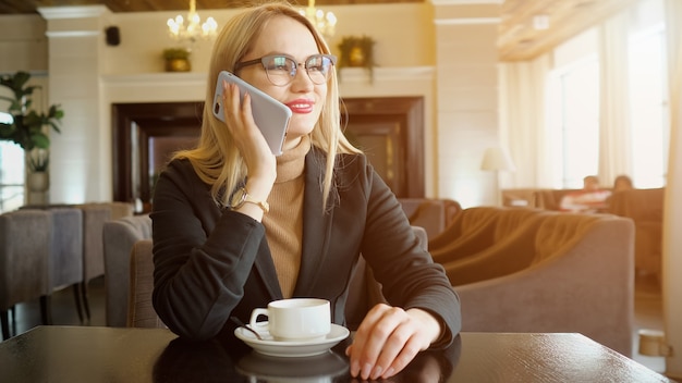 Sorridente giovane donna che ha una conversazione telefonica amichevole mentre beve caffè al bar, luce solare