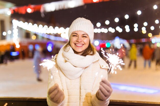 Sorridente giovane donna che indossa abiti invernali a maglia che tengono sparkler all'aperto su sfondo di neve