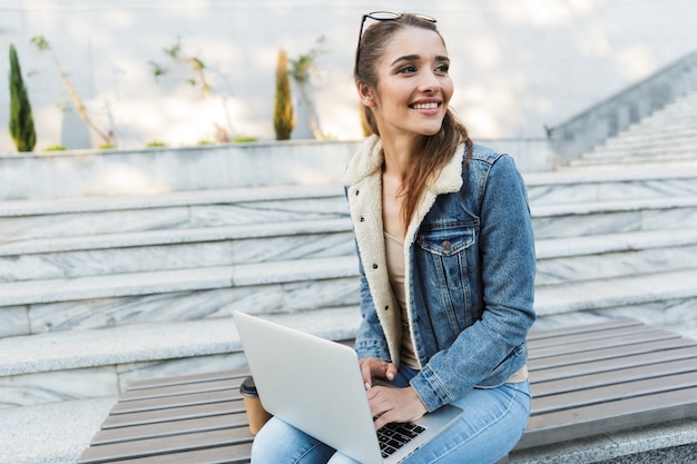 Sorridente giovane donna che indossa giacca seduta su una panchina all'aperto, utilizzando il computer portatile