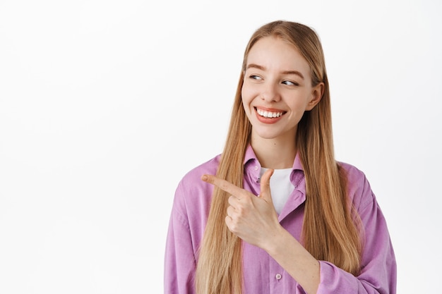 Sorridente giovane donna che sembra felice, puntando le dita e fissando da parte lo spazio della copia, in piedi contro il muro bianco