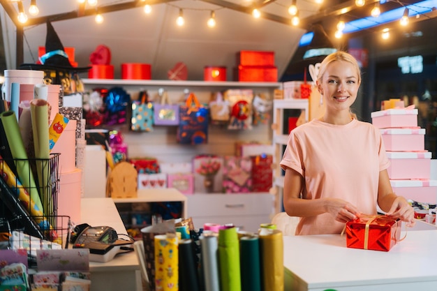 Sorridente giovane donna che tiene e usa un nastro dorato per legare l'arco per una confezione regalo avvolta in rosso per