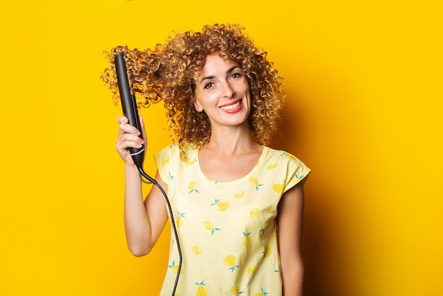 Sorridente giovane donna con i capelli ricci raddrizza i capelli con una piastra per capelli su uno sfondo giallo.