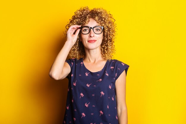 Sorridente giovane donna dai capelli ricci in bicchieri trasparenti su sfondo giallo.