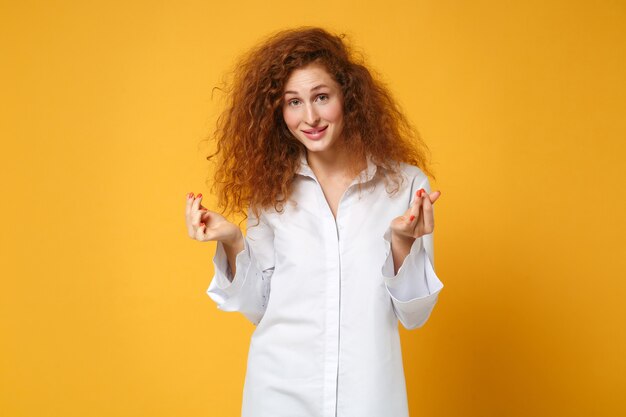 Sorridente giovane donna dai capelli rossi in camicia bianca casual in posa isolata sul muro giallo arancione