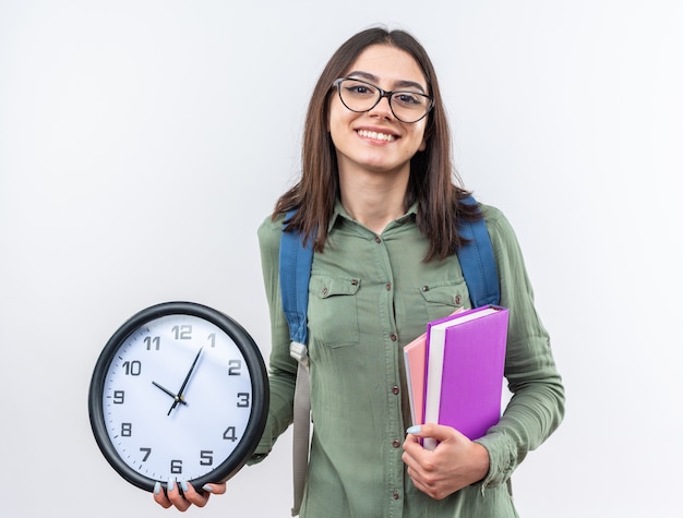 Sorridente giovane donna della scuola con gli occhiali con zaino in possesso di libri con orologio da parete