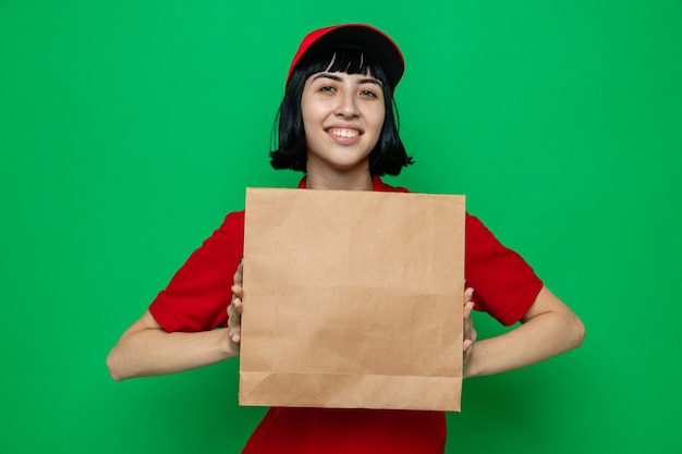 Sorridente giovane donna di consegna caucasica che tiene in mano un sacchetto di carta per alimenti e guarda