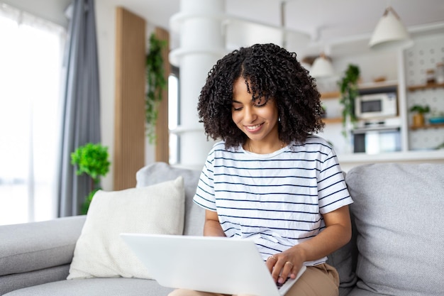 Sorridente giovane donna freelance che lavora da casa seduto sul divano utilizzando lo spazio della copia del laptop Rilassata signora afroamericana che si gode il suo fine settimana navigando su Internet sul laptop