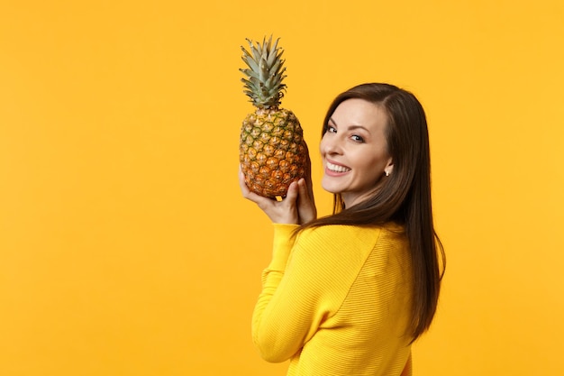 Sorridente giovane donna in abiti casual guardando indietro, tenendo frutta fresca di ananas maturo isolato su sfondo giallo muro arancione. Persone stile di vita vivido, relax concetto di vacanza. Mock up spazio di copia.