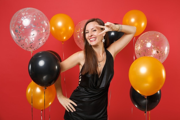 Sorridente giovane donna in abito nero che celebra, mostrando il segno della vittoria su palloni ad aria di sfondo rosso brillante. Concetto di festa di compleanno di mockup di compleanno di San Valentino, Giornata internazionale della donna felice anno nuovo.