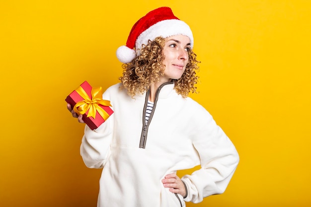 Sorridente giovane donna in cappello di Babbo Natale con confezione regalo su sfondo giallo.