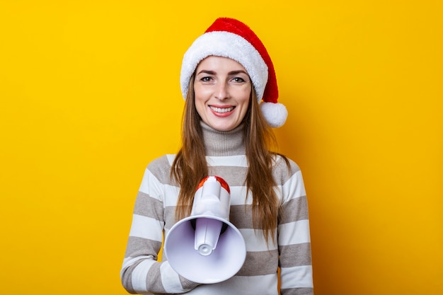 Sorridente giovane donna in cappello di Babbo Natale con un megafono su sfondo giallo.