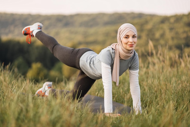 Sorridente giovane donna in hijab e abbigliamento sportivo facendo esercizi flessibili sul tappetino yoga