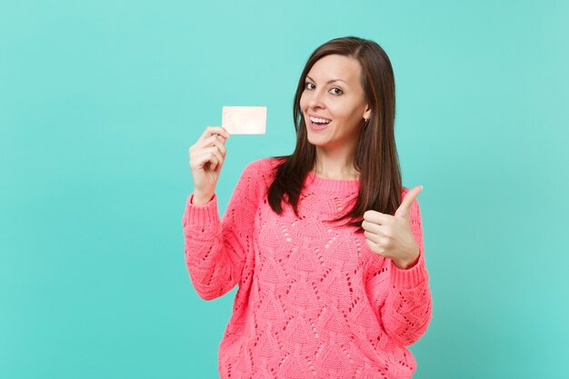 Sorridente giovane donna in maglia maglione rosa che mostra il pollice in alto e tenendo la carta di credito in mano isolato su blu turchese parete sfondo ritratto in studio. Concetto di stile di vita della gente. Mock up copia spazio.