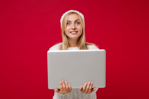 Sorridente giovane donna in maglione bianco cappello isolato su sfondo muro rosso in studio. Persone di stile di vita di moda sano emozioni sincere, concetto di stagione fredda. Simula lo spazio della copia. Tenere il computer portatile.