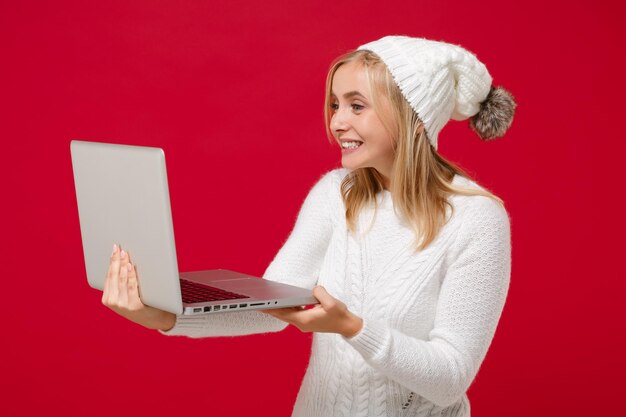 Sorridente giovane donna in maglione bianco cappello isolato su sfondo muro rosso in studio. Persone di stile di vita di moda sano emozioni sincere, concetto di stagione fredda. Simula lo spazio della copia. Tenere il computer portatile.