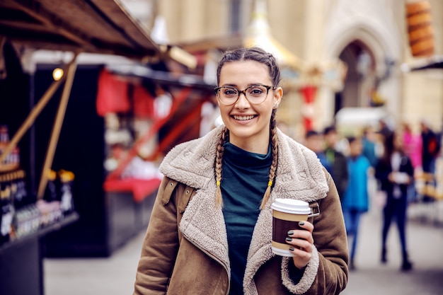 Sorridente giovane donna in piedi sulla strada il clima freddo e tenendo la tazza usa e getta con caffè.