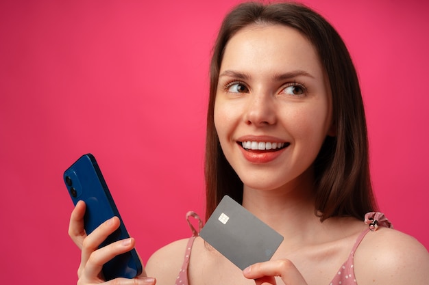 Sorridente giovane donna in possesso di smartphone e carta di credito contro backgorund rosa pink