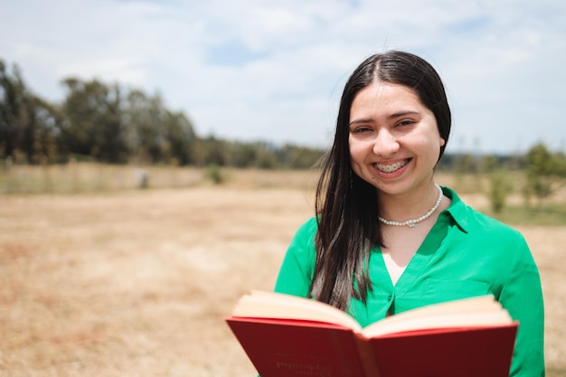 Sorridente giovane donna in possesso di un libro aperto nel campo e guardando la fotocamera Benefici della lettura
