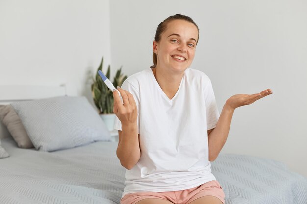 Sorridente giovane donna incinta adulta con test di gravidanza in posa nella sua camera da letto, indossando una maglietta bianca, scrollando le spalle, positivamente stupita di avere tali notizie.