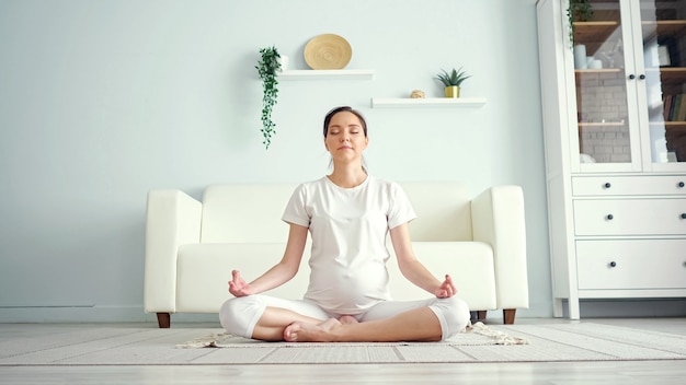 Sorridente giovane donna incinta in bianco medita seduto in posizione yoga padmasana sul pavimento contro mobili di design in una spaziosa stanza a casa