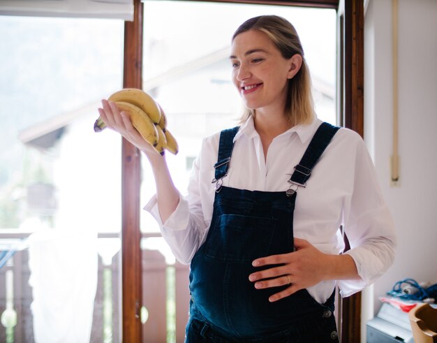 Sorridente giovane donna incinta in piedi e guardando un grappolo di banane fresche