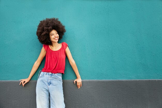 Sorridente giovane donna marocchina carina con acconciatura afro che distoglie lo sguardo mentre sta in piedi appoggiata al muro blu con le mani sulla superficie grigia alla luce del giorno