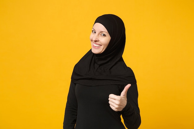 Sorridente giovane donna musulmana araba in abiti neri hijab che mostra i pollici in su, isolata sulla parete gialla, ritratto. Concetto di stile di vita religioso della gente.