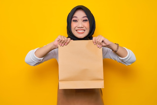 Sorridente giovane donna musulmana asiatica barista in grembiule maglietta bianca lavoro in caffetteria tenere carta bianca borsa da asporto mock up isolato su sfondo giallo Avvio aziendale