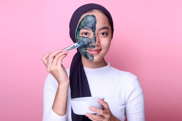 Sorridente giovane donna musulmana asiatica che applica l'argilla curativa cosmetica della maschera di spirulina a metà faccia usando