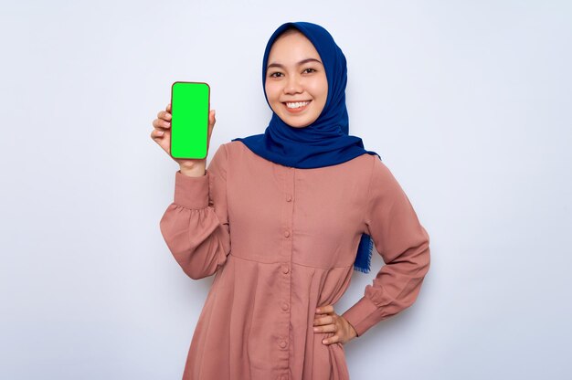 Sorridente giovane donna musulmana asiatica in camicia rosa che mostra lo schermo vuoto del telefono cellulare che consiglia App isolata su sfondo bianco Concetto di stile di vita religioso della gente