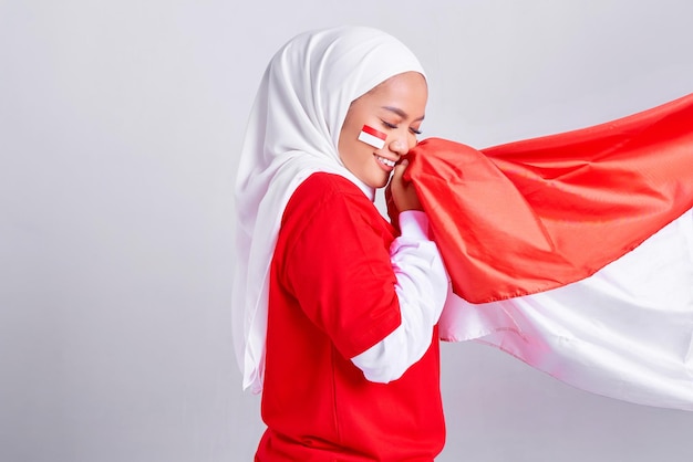 Sorridente giovane donna musulmana asiatica in maglietta bianca rossa rispetta o saluta gesto mentre bacia bandiera indonesiana e celebra il giorno dell'indipendenza indonesiana 17 agosto isolato su sfondo bianco