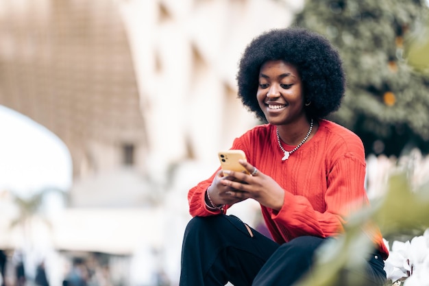 Sorridente giovane donna nera con l'acconciatura afro utilizzando il suo smartphone per strada