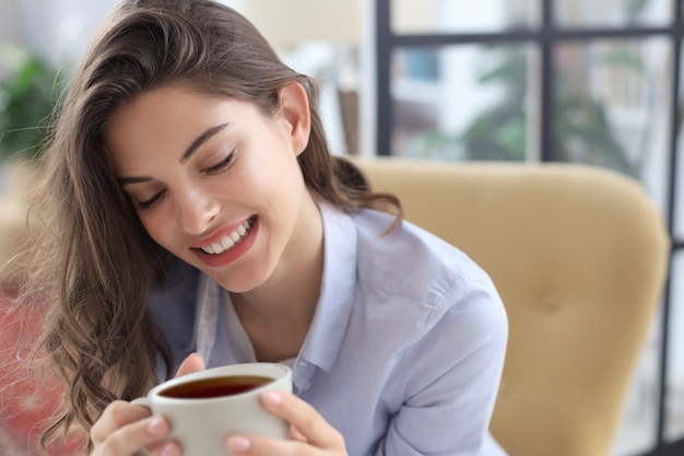 Sorridente giovane donna seduta in poltrona in soggiorno, con in mano una tazza di caffè.