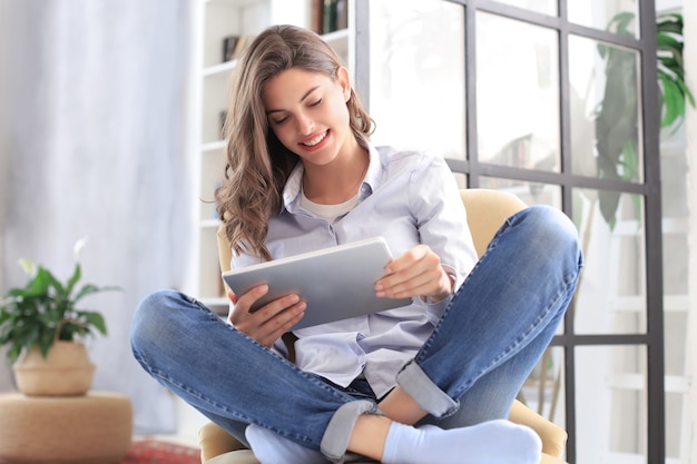 Sorridente giovane donna seduta in poltrona in soggiorno e utilizzando con il suo tablet.