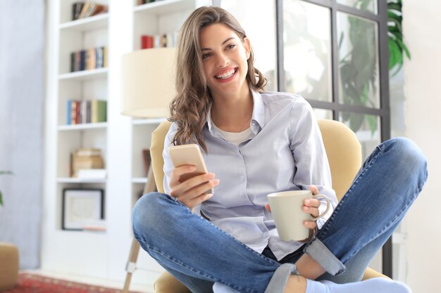 Sorridente giovane donna seduta in poltrona in soggiorno e utilizzando con il suo telefono cellulare.