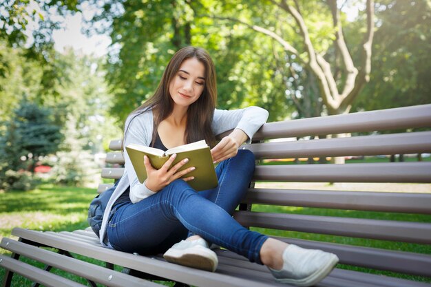 Sorridente giovane donna seduta su una panchina nel parco. Ragazza castana che si rilassa e che legge un libro. Stile di vita e concetto di refrigerazione