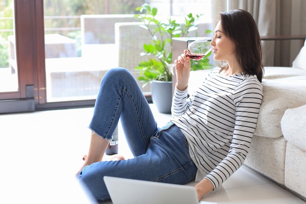 Sorridente giovane donna seduta sul pavimento con il computer portatile e chiacchierando con gli amici, bevendo vino.