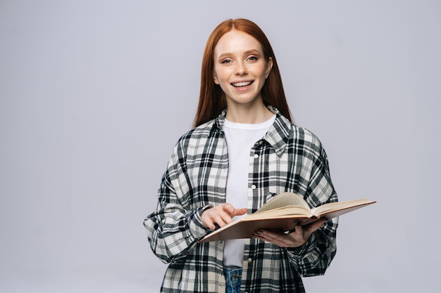 Sorridente giovane donna studente di college che gira le pagine del libro mentre legge e guarda la fotocamera su sfondo grigio isolato Modello di signora piuttosto rossa che mostra emotivamente le espressioni facciali