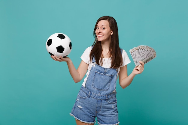 Sorridente giovane donna tifoso di calcio supporta la squadra preferita con pallone da calcio, fan di soldi in banconote in dollari, denaro contante isolato su sfondo blu turchese. Concetto di stile di vita per il tempo libero della famiglia sportiva.